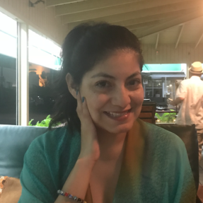 image of a women sitting in an outside restaurant with hair in ponytail and hand resting on side of face smiling.