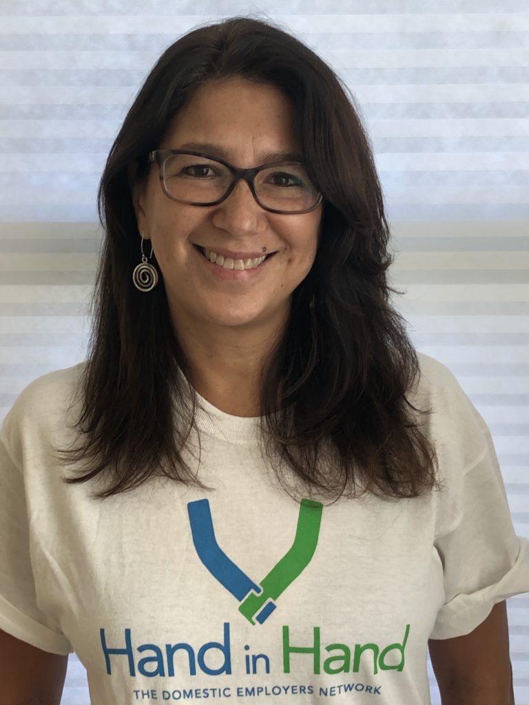 image of Latina woman with long brown hair and glasses smiling looking at the camera wearing a white t-shirt that reads Hand in Hand: The Domestic Employers Network