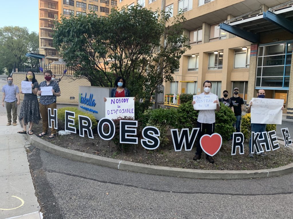 Photograph of people in masks standing 6 feet apart in front of a nursing home holding signs that read "Nobody is Disposable" Sign on the ground reads "Heroes Work Here"