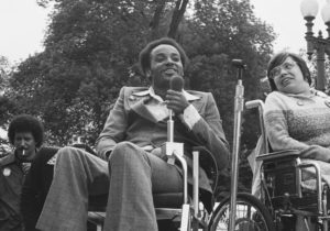 black and white image of black man wearing a suit in wheelchair speaking in microphone, sitting next to white woman in glasses in a wheelchair