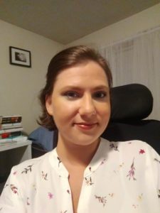 A white woman with short brown hair pinned back and smiling at the camera. She is wearing a white blouse with pink, red, yellow and green flowers. A black wheelchair headrest call be seen behind her head.