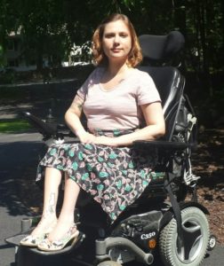 A white woman with short blonde hair is pictured outside. She is wearing a pink tee-shirt, black skirt with pink and green design, and matching heels. She is smiling toward the camera and seated in a black power wheelchair.