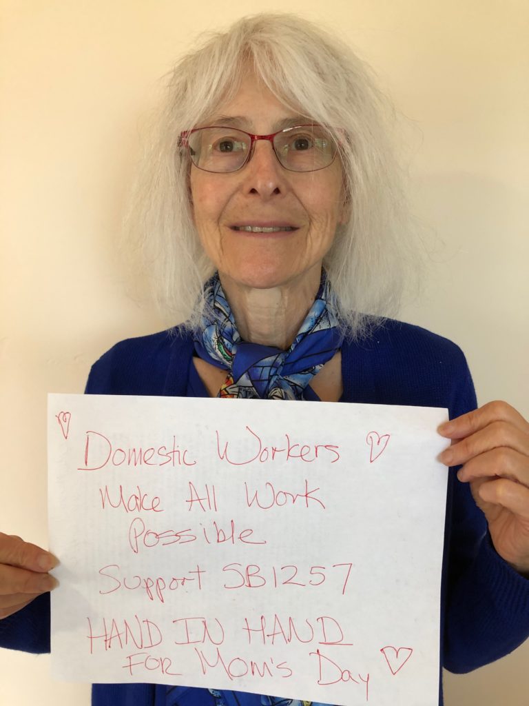 White woman with grey hair and glasses smiling holding a sign that reads "Domestic Workers make all work possible. Support SB1257"