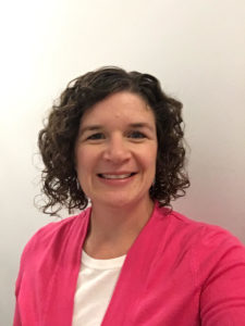 White woman with chin length curly brown hair smiles with pink sweater and white shirt