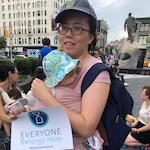 Asian woman wearing wearing gray hat, pink shirt, and glasses standing outside with infant to chest. 