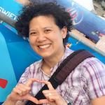 Woman with short black curly hair and plaid shirt standing outside making heart symbol with hands. 