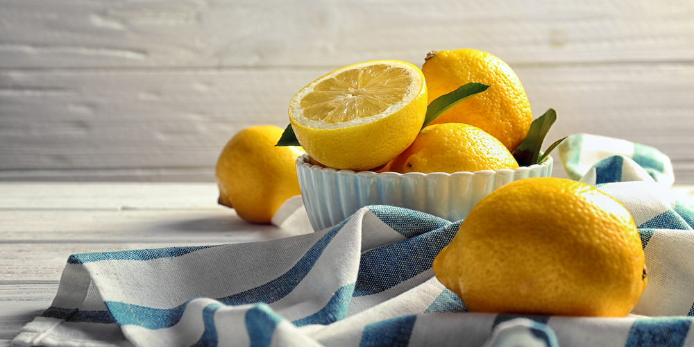 Halved and whole lemons sit on a counter in a white bowl and on a blue and white striped dishcloth