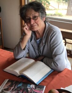 Image of white women with short grey hair with oxygen tube in her nose sitting in front of a book looking at the camera and gently smiling.