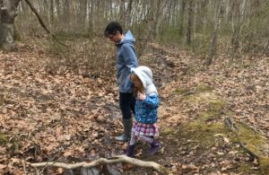 image of white woman wearing rain jacket and boots holding hand with young child in a jacket, skirt and boots, walking in the woods