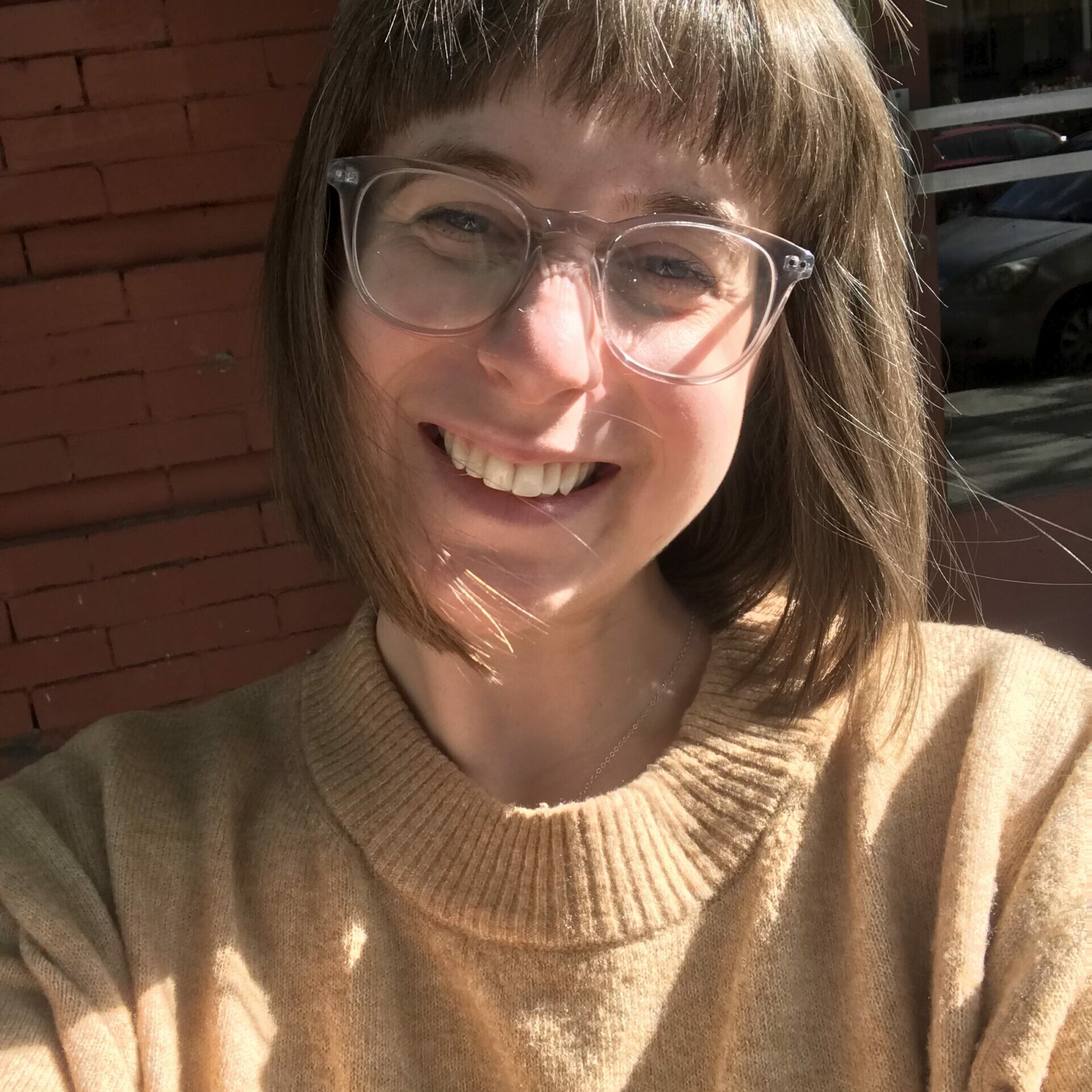 Image of white woman smiling with clear glasses and shoulder length light brown hair wearing a tan sweater. She is outside sunlights is hitting her face.