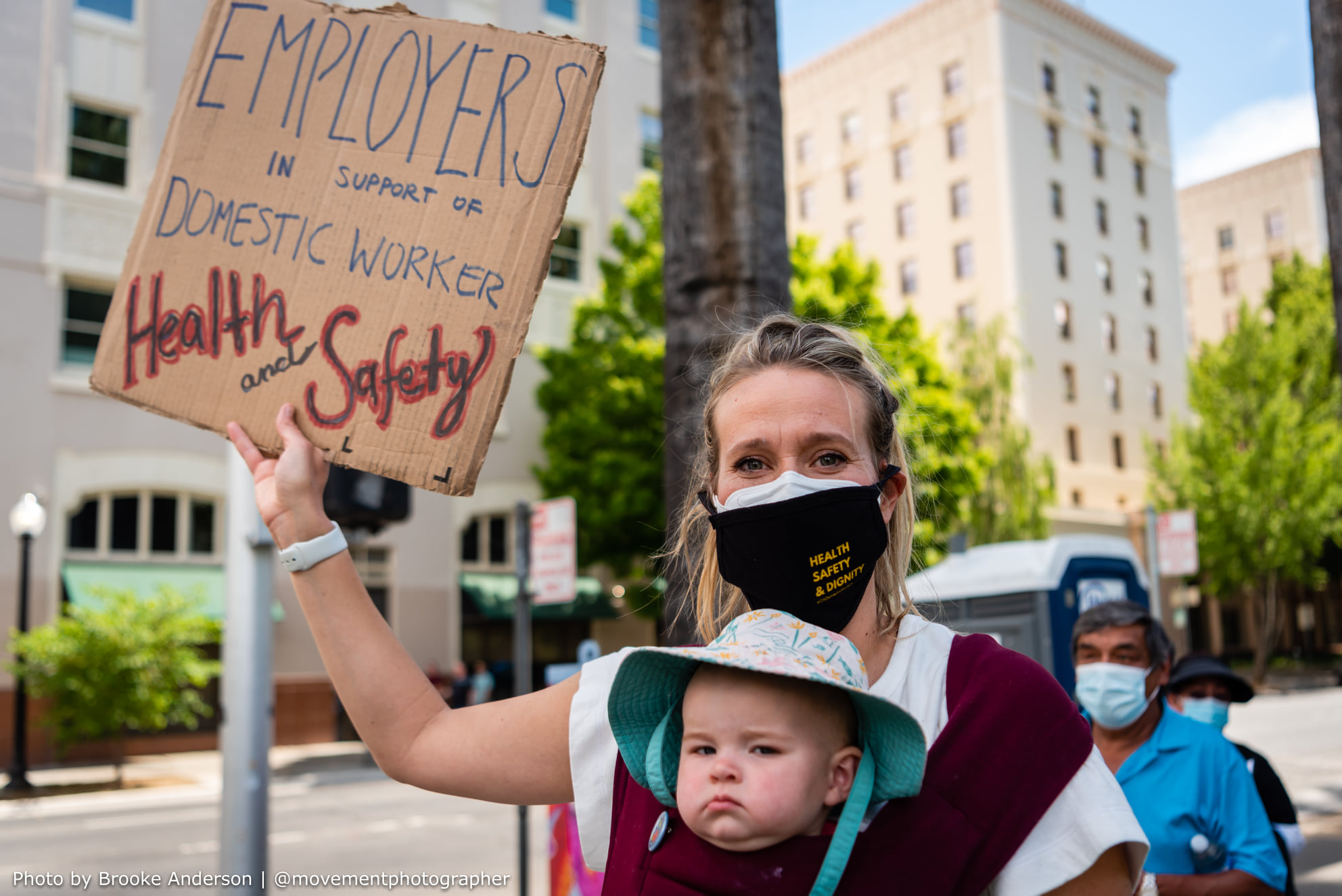 Woman holding baby and sign Employers in support of Domestic Worker Health and Safety. 