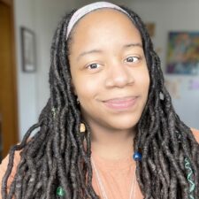 Image of black woman with long dreadlocks smiling and looking directly in the camera. Wearing a peach colored shirt and silver necklace.