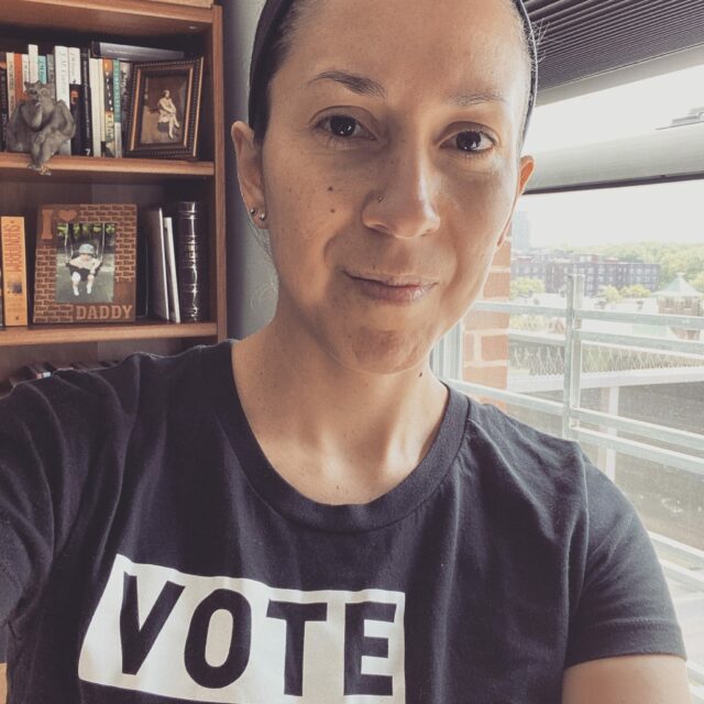 image of Latina woman softly smiling hair pulled back wearing a t-shirt that reads "vote like a mother"
