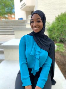 A young black woman who is wearing a black hijab, blue blouse, and black pants is sitting outside.