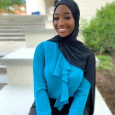 A young black woman who is wearing a black hijab, blue blouse, and black pants is sitting outside.