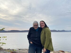 Two white women stand in front of a lake as the sun starts to set.