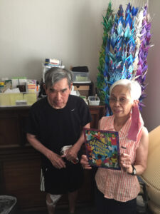 An elderly couple stand next to each other holding the Honor Domestic Work poster