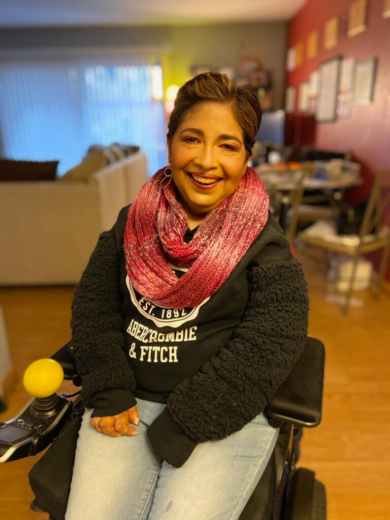 Woman with tan skin, short brown hair with blond streaks smiles at the camera from a power wheelchair, wearing a pink scarf and dark gray sweatshirt. A blurred background appears to be a living room with red walls covered in photos , a small table and white coach