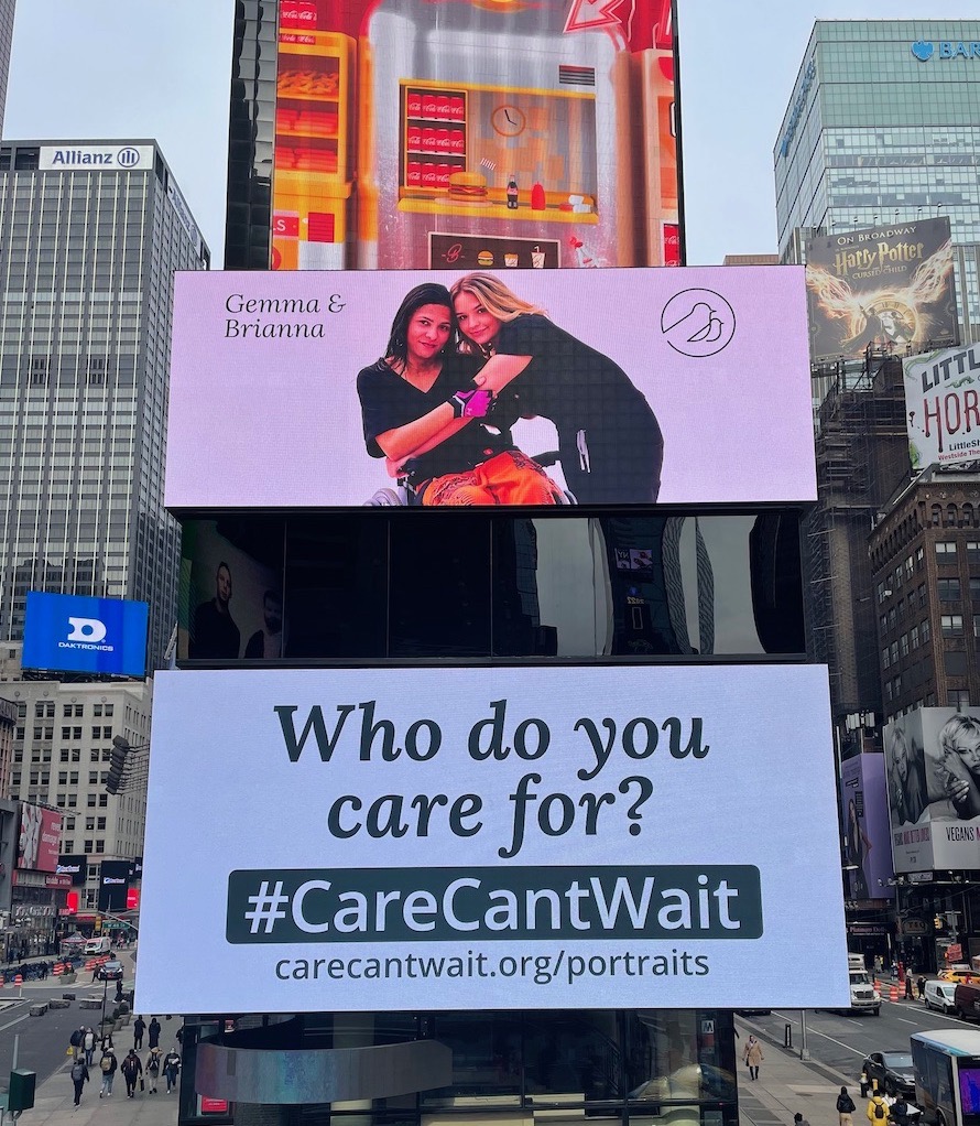 Billboard image in middle of busy city, with image of woman in wheelchair with black hair hugging woman to her right with blonde hair