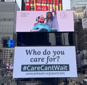 billboard in Times Square. Photograph shows white man in wheelchair next to a black man with his arm around him. Text reads Who do you care for #CareCantWait