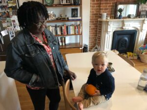 Black women standing looking a white baby on table holding a pumpkin 