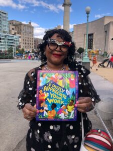 Photo of a black women with beautifully decorated red and black glasses frame standing outside holding Honor Domestic Work poster