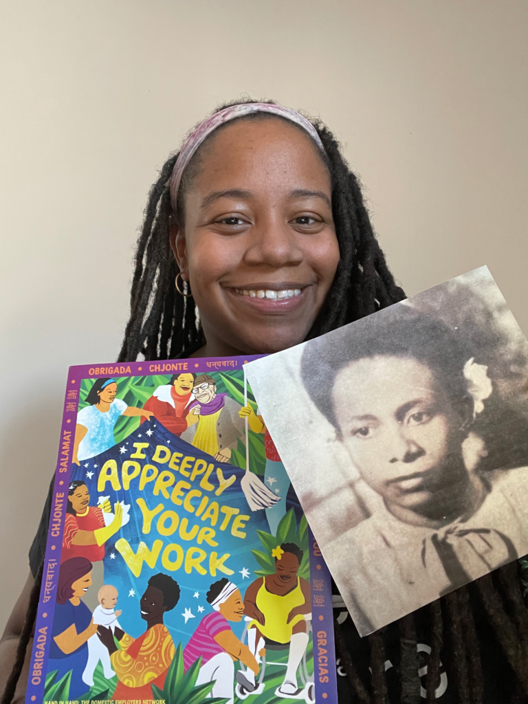 A black woman with locs smiling directly into camera against wall, holding Honor Domestic Work poster in her left hand and a black and white photo of her grandmother in her right hand.