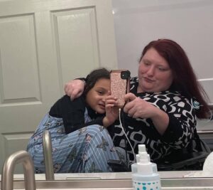 White woman in a wheelchair takes a selfie photo in the mirror with a long girl on her lap hugging her