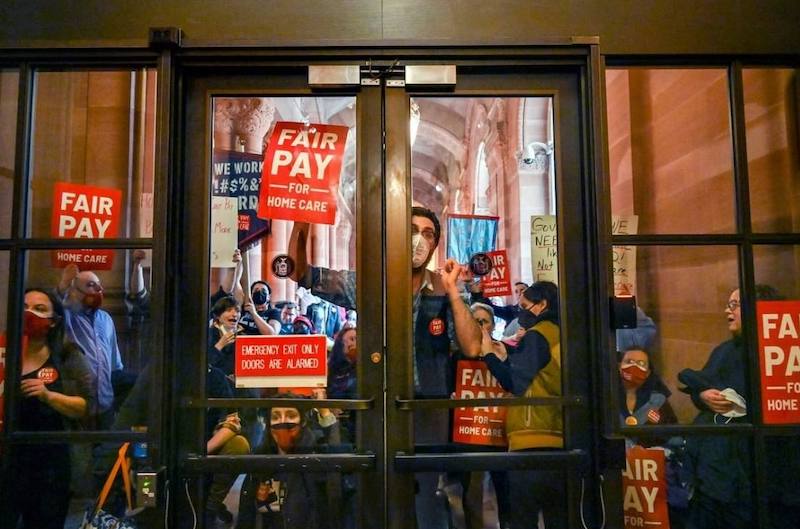 A crowd of people behind glass doors, pressing signs against the doors that say "Fair Pay for Home Care"