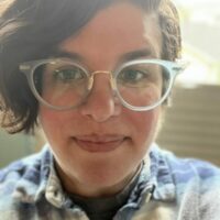 Headshot of white woman with brown hair wearing glasses and a plaid blue and white shirt, looking directly into the camera.