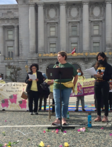 photo of Gretchen Sandler wearing dark green shirt and jeans standing in front of a large building outside speaking at a podium.