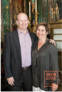 photo of a white man in a suit on the left and a white woman in a grey dress on the right smiling while the man has his hand on the woman's shoulder.