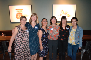 A photo of six women lined together smiling for a photo in front of two paintings on the wall.