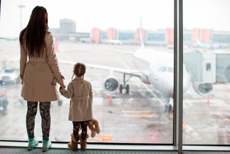 Woman and child hold hands in an airport staring out a large window looking at planes