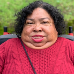 Headshot of black woman with short curly brown hair and red shirt wearing red lipstick is sitting in a wheelchair outside smiling.