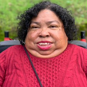 Headshot of black woman with short curly brown hair and red shirt wearining red lipstick is sitting in wheelchair outside smiling.