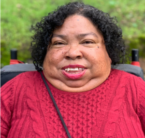 Headshot of black woman with short curly brown hair and red shirt wearining red lipstick is sitting in wheelchair outside smiling.