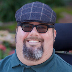 White man with beard sitting in a wheelchair wearing black sunglasses, hat, and a dark green shirt.