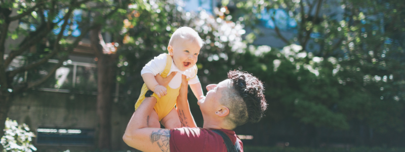 parent with short curly mohawk holding baby wearing yellow overalls up towards the sky, as the baby smiles outside. 