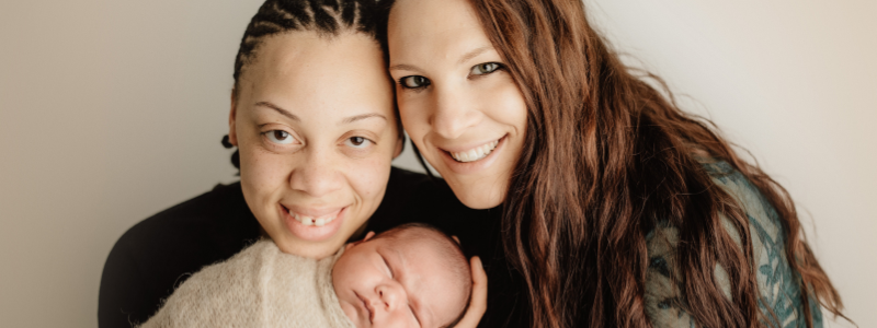 family portrait of two moms smiling towards the camera while the mom on the left holds the infant that snuggled in a tan blanket.