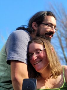 sage and her partner Mark outside. Sage is smilling while her head gently rest on Mark's arm. Mark is standing above sage looking in the opposite direction. He is wearing glasses and a blue t-shirt. 