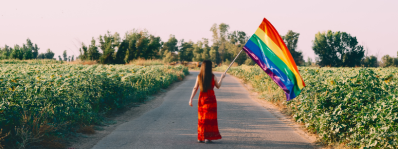 a person standing outside with back towards the camera wearing a red dress while holding the pride flag.