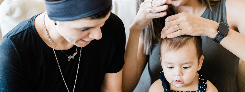 Photo of baby getting hair put in pigtails while a person to the left of her with black shirt and black hat smiles at baby.