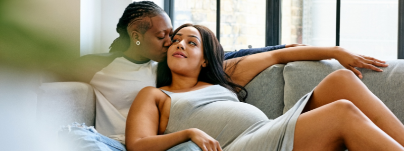 A couple laying on a couch a person on the left who is black with braided hair kisses the person on the right who has long brown hair as she reclines on her lap.