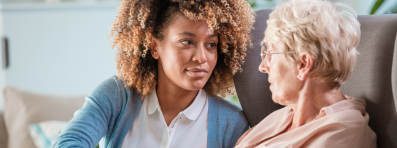 two people seated on a sofa. A Black woman in a white blouse with a blue sweater is speaking to an older white woman in a peach top.