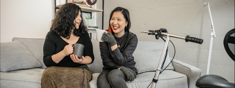 Two women, a white woman and an Asian woman, sitting on a sofa drinking coffee and talking joyously. The Asian woman is seated near a scooter mobility device.