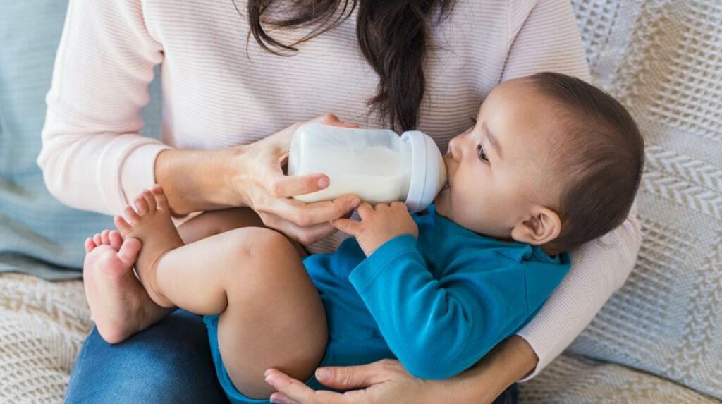 White woman holding baby with blue onesie while feeding baby a bottle.