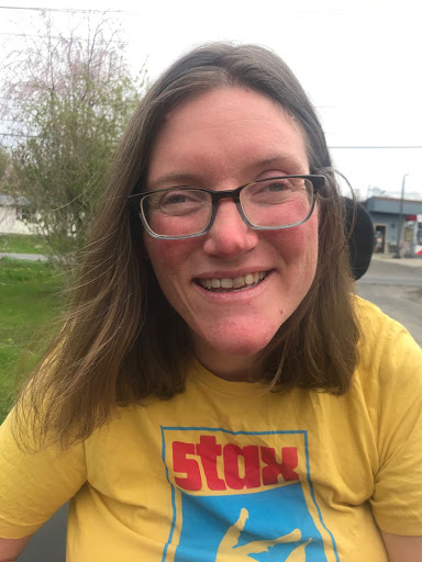 a white woman sitting in wheel chair outside smiling into camera while wearing glasses and a yellow shirt.
