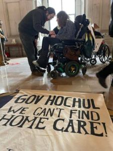 sage jobsis seated in wheel chair in front of large scrool with text "Gov Hochul we can't find Home Care!" as her partner bends down slightly in front of her 