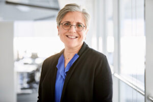 White woman with short gray hair and glasses wearing blue button up shirt and black blazer smiling at camera.